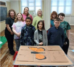 5 students and 4 teachers smile for a picture in front of a butterfly painted on cardboard.