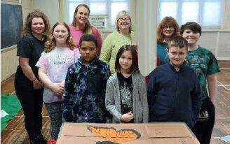5 students and 4 teachers smile for a picture in front of a butterfly painted on cardboard.