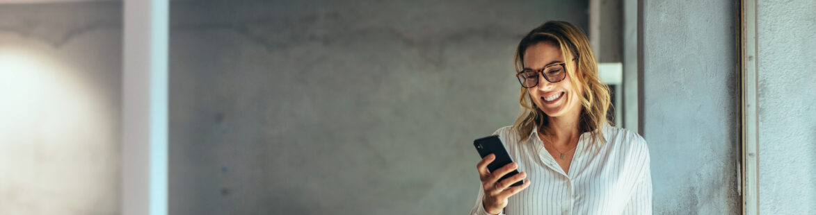 Smiling businesswoman using her phone in the office. Small business entrepreneur looking at her mobile phone and smiling while communicating with her office colleagues