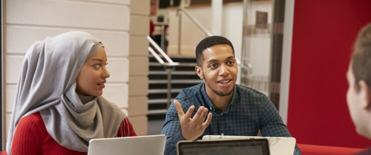 Photo of three people at a table with laptops. One man is facing another man, and a woman. The man next to the woman is talking and the other two are looking at him.