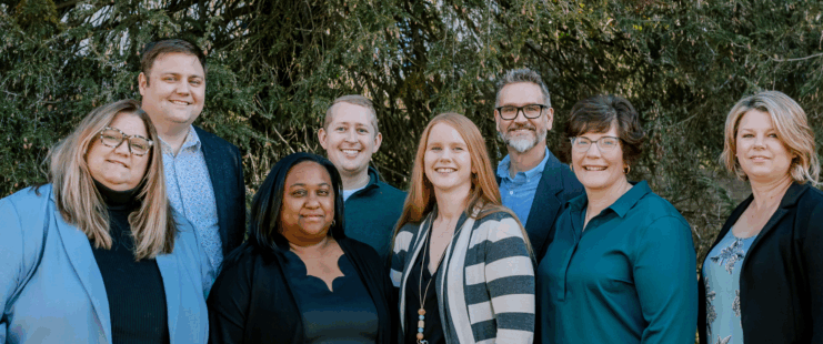 Group of eight adults smiling and standing in front of background greenery.