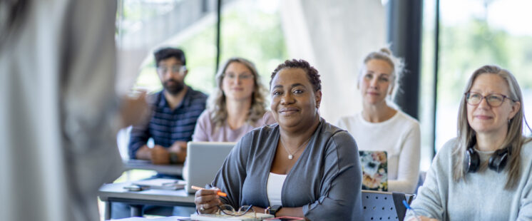 A large group of diverse mature students, sit together in rows as they listen attentively to their teacher explain the lesson. They are each dressed casually and have notebooks, textbooks, laptops and papers out in front of them for reference and taking notes.