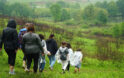 A group of adults and children walk downhill through a green, grassy field on a rainy day, some wearing clear ponchos, with trees and a pond visible in the background.
