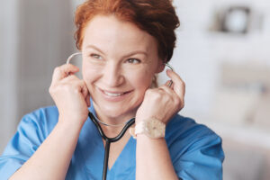 Healthy patient. Radiant medical worker looking into vacancy and grinning widely while standing with a stethoscope and listening to a patients heart beating.