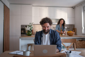 Bearded man smiling, working on a laptop at a kitchen table, while a woman in the background is happily preparing food in a bright, modern home kitchen